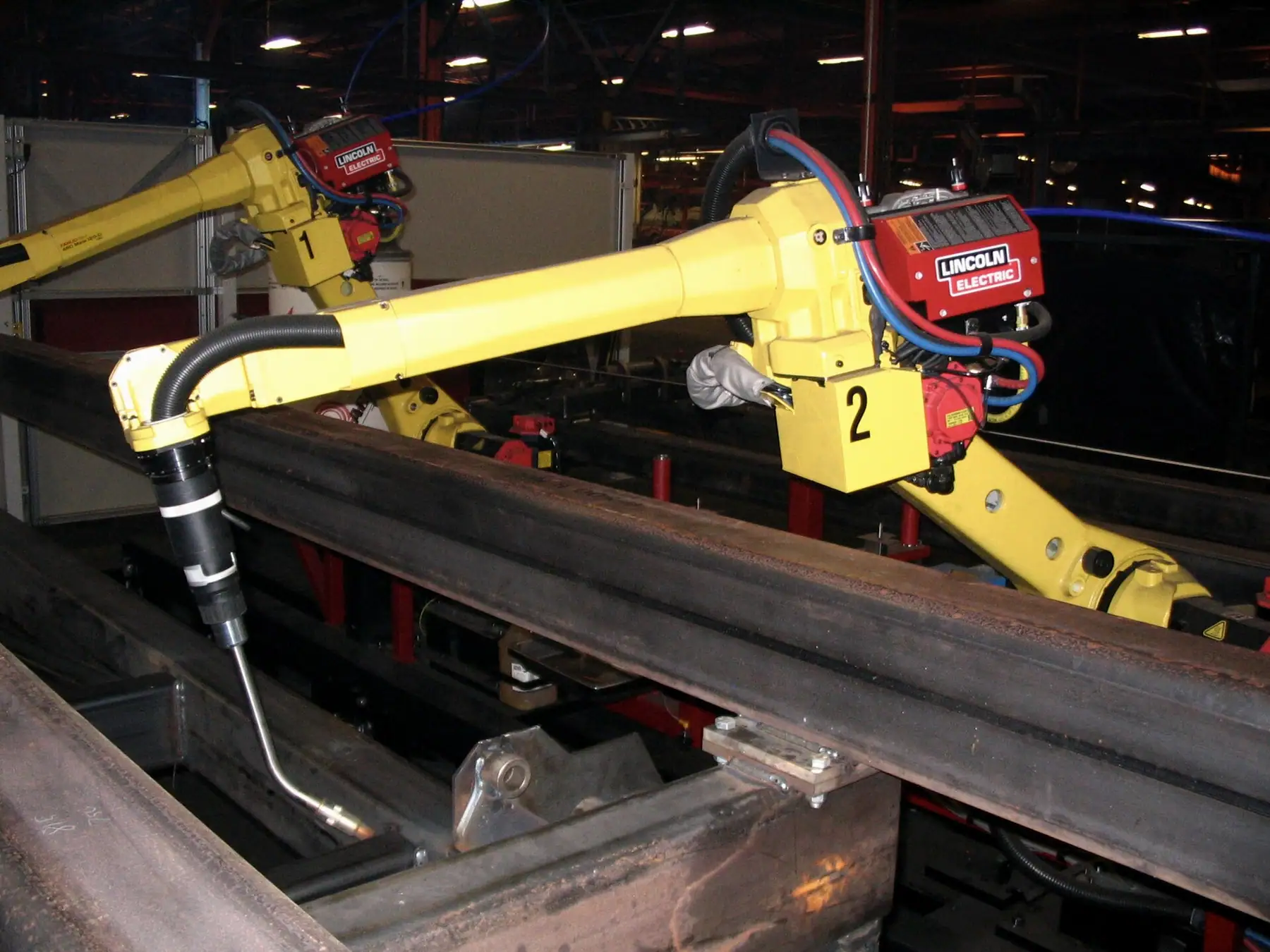Kaynak torcuyla uzanan sarı robot kolu ve arka planda kaynak güç kaynağı
